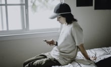 A queer teenager looks at their phone while sitting on a bed.