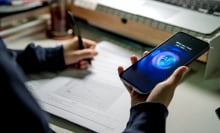 A teen holds a phone in their hand and consults an AI for help while writing in a notebook. 