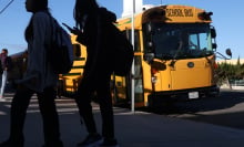 Students holding cellphones walk by a yellow school bus.
