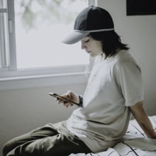 A queer teenager looks at their phone while sitting on a bed.