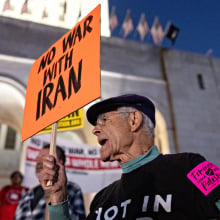 A person stands amid a crowd, holding an orange protest sign that reads "No war with Iran."