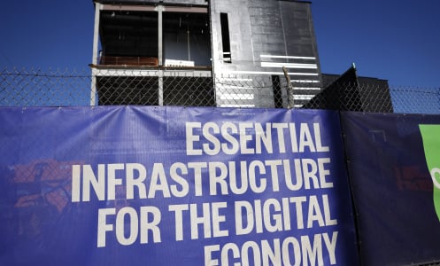 A large data center building under construction. A sign in front reads "essential infrastructure for the digital economy."