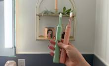 Hand holding pink and green Suri toothbrushes in front of bathroom wall