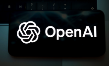 The OpenAI logo appears on the screen of a smartphone placed on a laptop keyboard illuminated by green light.